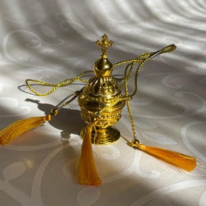 May include: A gold-colored metal censer with a cross on top and a chain with tassels. The censer is used in religious ceremonies to burn incense.