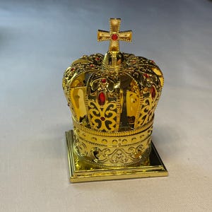 以下が含まれることがあります： 赤い宝石がちりばめられた、十字架の付いた金色の王冠。王冠は正方形の台座にあり、複雑な透かし彫りと装飾模様が特徴です。威厳のある華やかな装飾品。