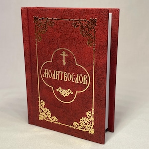 May include: A red book with gold embossed lettering and decorative flourishes. The book's cover features the word "MOLITVOSLOV" in gold lettering, with a cross above it. The book is slightly open, revealing white pages.