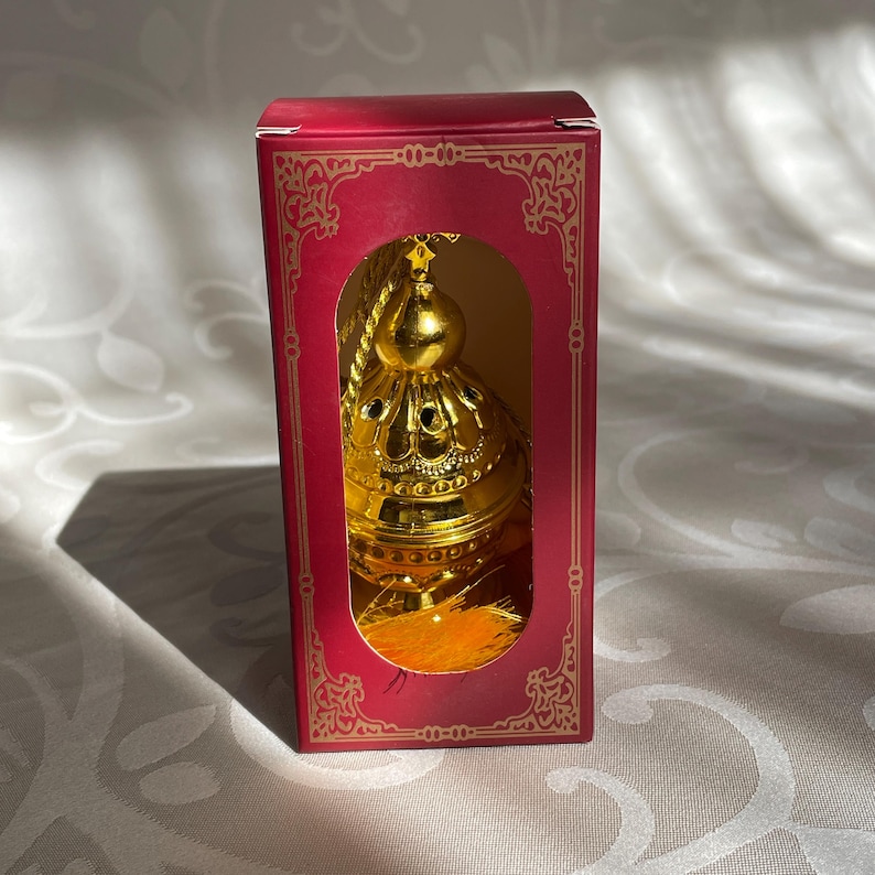May include: A gold metal incense burner with a decorative design and a tassel. The burner is in a red box with gold trim.