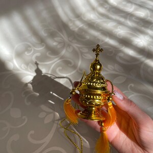May include: A gold censer with a cross on top and a chain. The censer has a round body with intricate details and is decorated with small beads. It has two yellow tassels hanging from the sides.