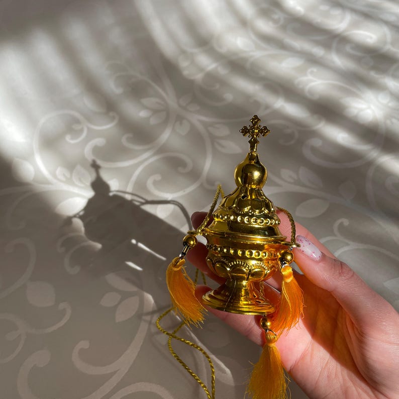 May include: A gold censer with a cross on top and a chain. The censer has a round body with intricate details and is decorated with small beads. It has two yellow tassels hanging from the sides.