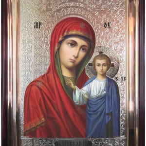May include: A religious icon featuring a woman in a red head covering and robe, with a child in a blue robe. The icon is set in a dark brown wooden frame with a silver background and text at the bottom.