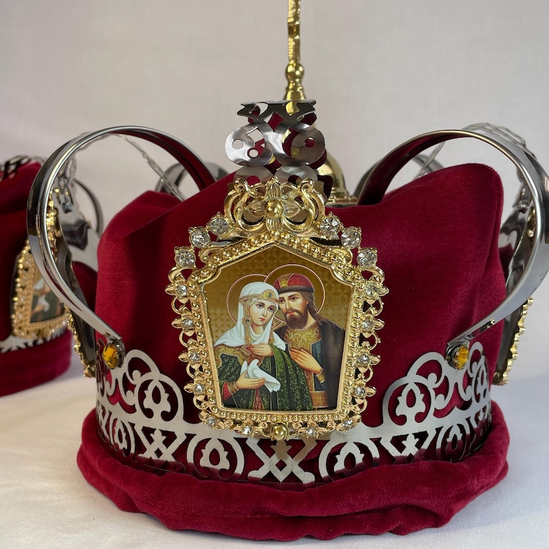May include: A decorative crown with a deep red velvet base, adorned with silver and gold accents. The crown features an ornate gold frame with a religious icon and sparkling embellishments. The design includes intricate metalwork and a tall, decorative finial.