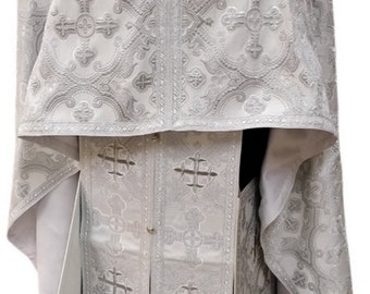 May include: A white liturgical vestment with intricate silver embroidery featuring crosses and floral patterns. The vestment has a long, flowing design with a high collar and a front closure.