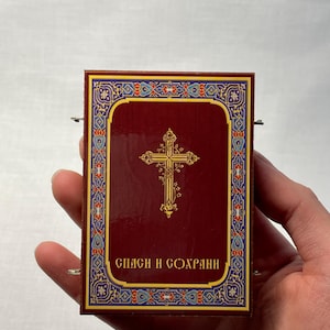 K&ouml;nnte beinhalten: Eine kleine, rechteckige Schachtel in Rot mit einem goldenen Kreuz und einem verzierten Rand. Die Schachtel tr&auml;gt den Text "СПАСИ И СОХРАНИ" in goldenen Buchstaben.