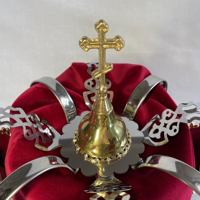 May include: A gold-colored religious crown with a cross on top, set against a deep red velvet cushion. The crown features silver metal bands and decorative silver filigree elements, creating a regal appearance.