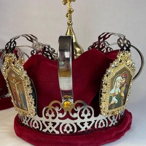May include: A decorative Orthodox Christian wedding crown. The crown features a silver filigree design with a red velvet interior. Two gold-framed icons with religious imagery are attached. A gold cross sits atop the crown.