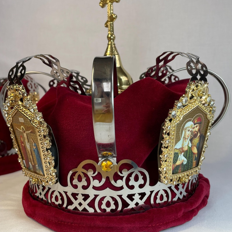 May include: A decorative Orthodox Christian wedding crown. The crown features a silver filigree design with a red velvet interior. Two gold-framed icons with religious imagery are attached. A gold cross sits atop the crown.