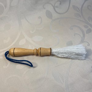 Puede incluir: Un cepillo de madera con un cabezal de cerdas blancas y un mango de cordón azul. El cepillo tiene un ribete de madera oscura alrededor del mango.