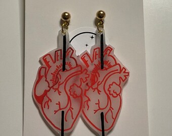 Könnte beinhalten: Ein Paar Ohrringe mit einem roten anatomischen Herzen, das von einem schwarzen Pfeil durchbohrt wird. Die Ohrringe sind aus klarem Acryl und haben goldfarbene Haken.