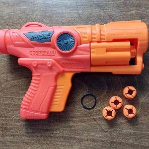 May include: A red and orange toy gun with the words "Adventure Force" and "Desperado" printed on the side. The gun comes with five orange plastic projectiles.