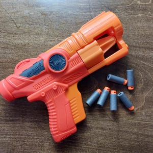 Op de afbeelding: Een oranje en zwart speelgoedpistool met de woorden "Ultimate Air Force" en "Desperado" erop gedrukt. Het pistool is omringd door blauwe en oranje foam darts.
