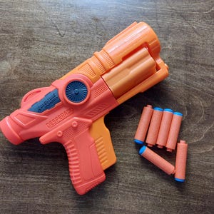 May include: An orange and black toy gun with the words "Wolverine Force" and "Desperado" printed on the side. The gun is accompanied by several orange and blue toy darts.