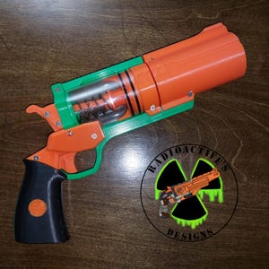 Puede incluir: Una pistola de juguete impresa en 3D en naranja, verde y negro. El arma tiene un mango, un gatillo y un cañón. La palabra "SILLY" es visible en el cañón. Un logotipo con el texto "Radioactive's Designs" está en la esquina inferior derecha.