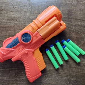 Puede incluir: Una pistola de juguete naranja y negra con las palabras "Full-Auto" y "DESPERADO" impresas en el lateral. La pistola va acompañada de seis dardos verdes y azules.