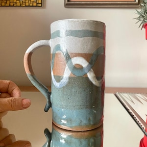 May include: A ceramic mug with a blue, white and brown abstract design. The mug has a handle and is made in a rustic style.