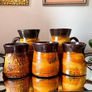 May include: Set of five brown and orange ceramic mugs with a speckled glaze. The mugs have a rounded shape and a handle on one side.