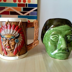Peut inclure: Deux tasses en céramique représentant des images amérindiennes. Une tasse est brune avec une représentation d'un homme amérindien portant une coiffe. L'autre tasse est verte et représente le visage d'un homme amérindien.