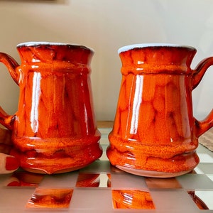 May include: Two orange ceramic mugs with white rims. The mugs have a textured, glazed finish and are slightly flared at the top.