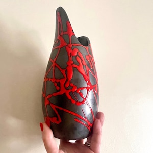 May include: A black ceramic vase with a red abstract design. The vase has a unique shape with a wide opening at the top and a narrow base.