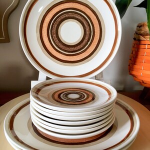 Op de afbeelding: Een set vintage keramische dinerborden met een bruin, oranje en wit concentrisch cirkelpatroon. De set bevat een groot dinerbord en een stapel kleinere borden.
