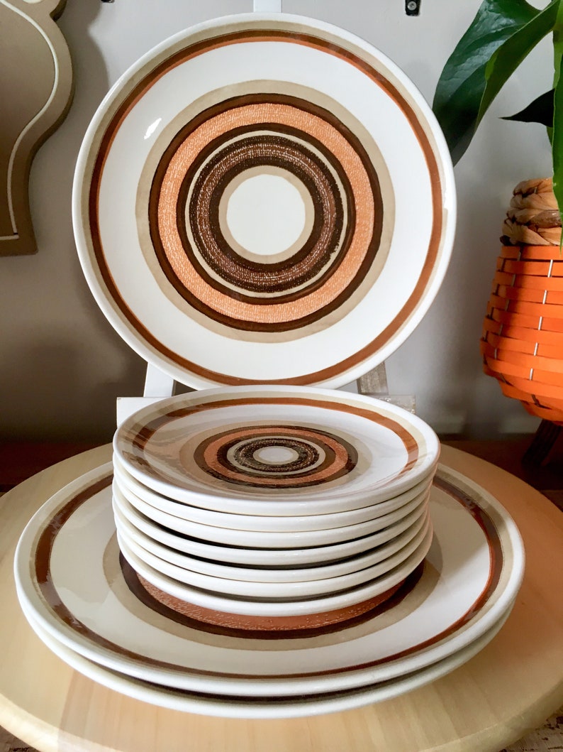 Op de afbeelding: Een set vintage keramische dinerborden met een bruin, oranje en wit concentrisch cirkelpatroon. De set bevat een groot dinerbord en een stapel kleinere borden.