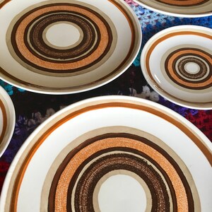 Op de afbeelding: Set van zes witte keramische borden met concentrische cirkels in bruin, oranje en beige. De borden zijn in een verspringend patroon op een kleurrijk gehaakt oppervlak gerangschikt.