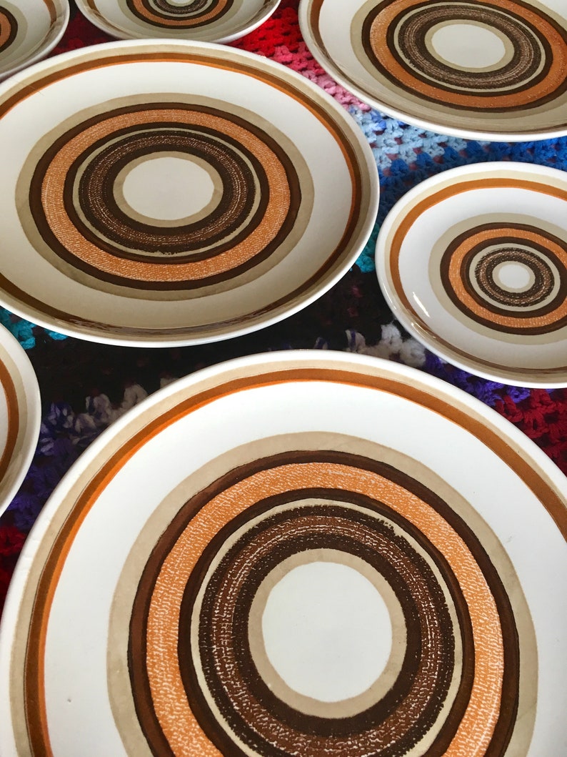 Op de afbeelding: Set van zes witte keramische borden met concentrische cirkels in bruin, oranje en beige. De borden zijn in een verspringend patroon op een kleurrijk gehaakt oppervlak gerangschikt.