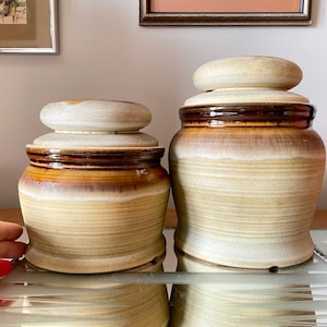 May include: Two ceramic jars with lids. The larger jar is brown and white with a light brown lid. The smaller jar is brown and white with a white lid. Both jars have a textured, striped design.