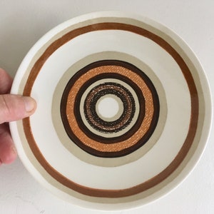 Op de afbeelding: Een witte keramische schotel met een concentrisch cirkelontwerp in tinten bruin, beige en oranje.