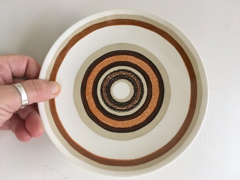 Op de afbeelding: Een witte keramische schotel met een concentrisch cirkelontwerp in tinten bruin, beige en oranje.