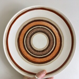 Op de afbeelding: Een wit keramiek dinerbord met een concentrisch cirkelontwerp in tinten bruin, oranje en beige.