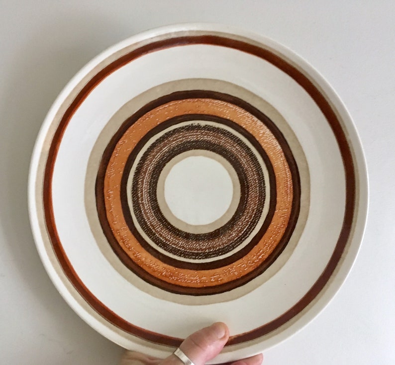 Op de afbeelding: Een wit keramiek dinerbord met een concentrisch cirkelontwerp in tinten bruin, oranje en beige.