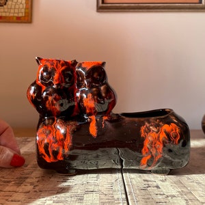 May include: A ceramic planter shaped like a log with two owls perched on top. The planter is decorated with a black and orange marbled glaze.