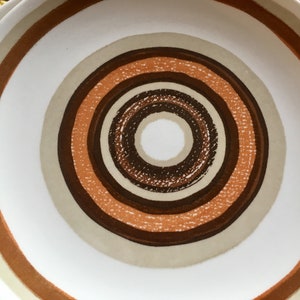 Op de afbeelding: Een wit keramiek bord met een concentrisch cirkelontwerp in tinten bruin, beige en oranje.
