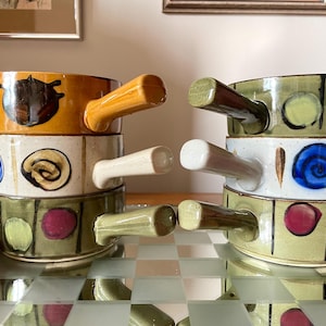 May include: Two sets of stacked ceramic bowls with handles. Each set has three bowls with different patterns. The top bowl in each set is yellow with a brown design. The middle bowl is white with a brown and blue design. The bottom bowl is green with brown and red dots.