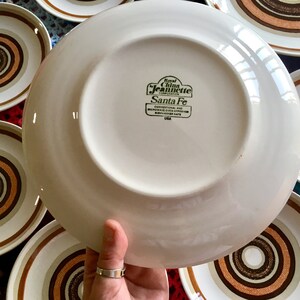 Op de afbeelding: Een wit keramiek bord met een bruin en wit concentrisch cirkelontwerp. Het bord heeft een groen label met de tekst "Royal China Jeanette Corporation Santa Fe Conventional and Microwave Oven Approved Dishwasher Safe USA".