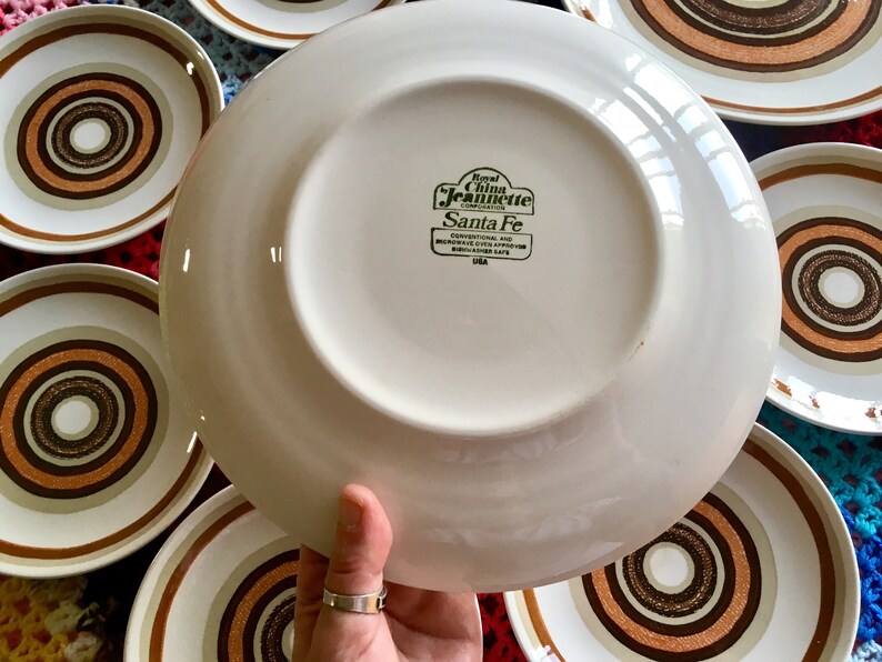 Op de afbeelding: Een wit keramiek bord met een bruin en wit concentrisch cirkelontwerp. Het bord heeft een groen label met de tekst "Royal China Jeanette Corporation Santa Fe Conventional and Microwave Oven Approved Dishwasher Safe USA".