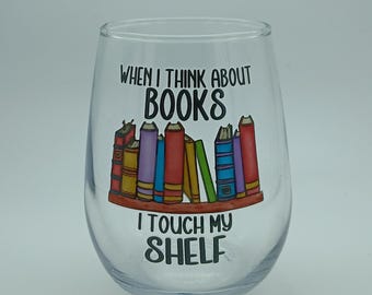 May include: A clear stemless wine glass with the text "WHEN I THINK ABOUT BOOKS I TOUCH MY SHELF." The glass features a colorful illustration of a bookshelf with various books in red, yellow, purple, and blue.