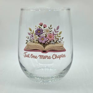 Op de afbeelding: Een helder glas zonder steel met een afbeelding van een open boek waaruit een boeket paarse, roze en witte bloemen groeit. Onder het boek staat de tekst "Just One More Chapter" gedrukt in een bruin cursief lettertype.