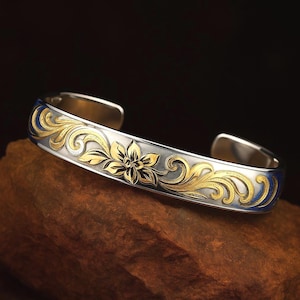 Puede incluir: Brazalete de plata con filigrana floral dorada y detalles de esmalte azul. El brazalete abierto presenta un diseño floral central y volutas ornamentadas. La joya se exhibe sobre una superficie texturizada marrón.