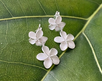 May include: A pair of silver earrings featuring a double plumeria flower design. The earrings are delicate and feminine, perfect for adding a touch of tropical elegance to any outfit.