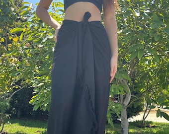 May include: A black sarong tied at the waist, paired with a matching tube top. The sarong has a fringed edge and is worn with white flip-flops. The scene is set outdoors with green grass and trees.