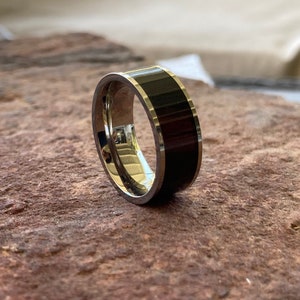 Puede incluir: Un anillo de plata con una incrustación de madera negra. El anillo está sobre una superficie marrón y texturizada.