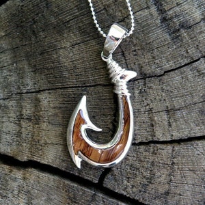 May include: A silver pendant necklace with a fish hook design. The hook has a wooden inlay and hangs from a silver chain. The pendant is set against a rustic wooden background.