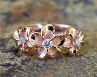 Puede incluir: Un anillo de oro rosa con tres diseños de flores de plumeria, cada uno con una pequeña piedra preciosa transparente en el centro.