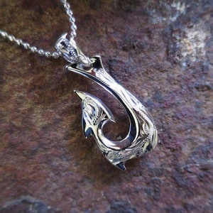 May include: A silver pendant necklace with a detailed fish hook design. The hook is intricately carved with floral patterns and hangs from a delicate chain. The pendant is set against a textured, brown background.