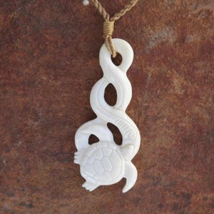 May include: A cream-coloured pendant necklace with a carved turtle design. The pendant is attached to a braided tan cord. The turtle has a detailed shell and head, and the pendant has an abstract, flowing shape.