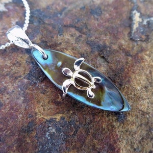 Pode incluir: Um colar de corrente de prata com um pingente que apresenta uma pequena tartaruga dourada em uma prancha de surf de abalone iridescente escura.